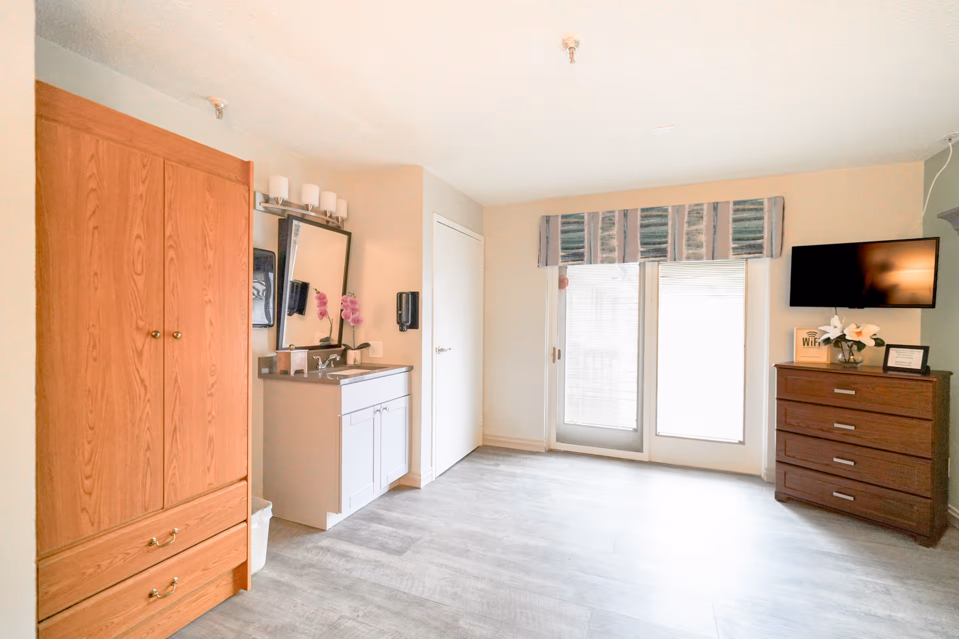 Bright private resident room with a wardrobe and vanity on the left, sliding glass doors ahead, and a dresser with a wall-mounted TV on the right.