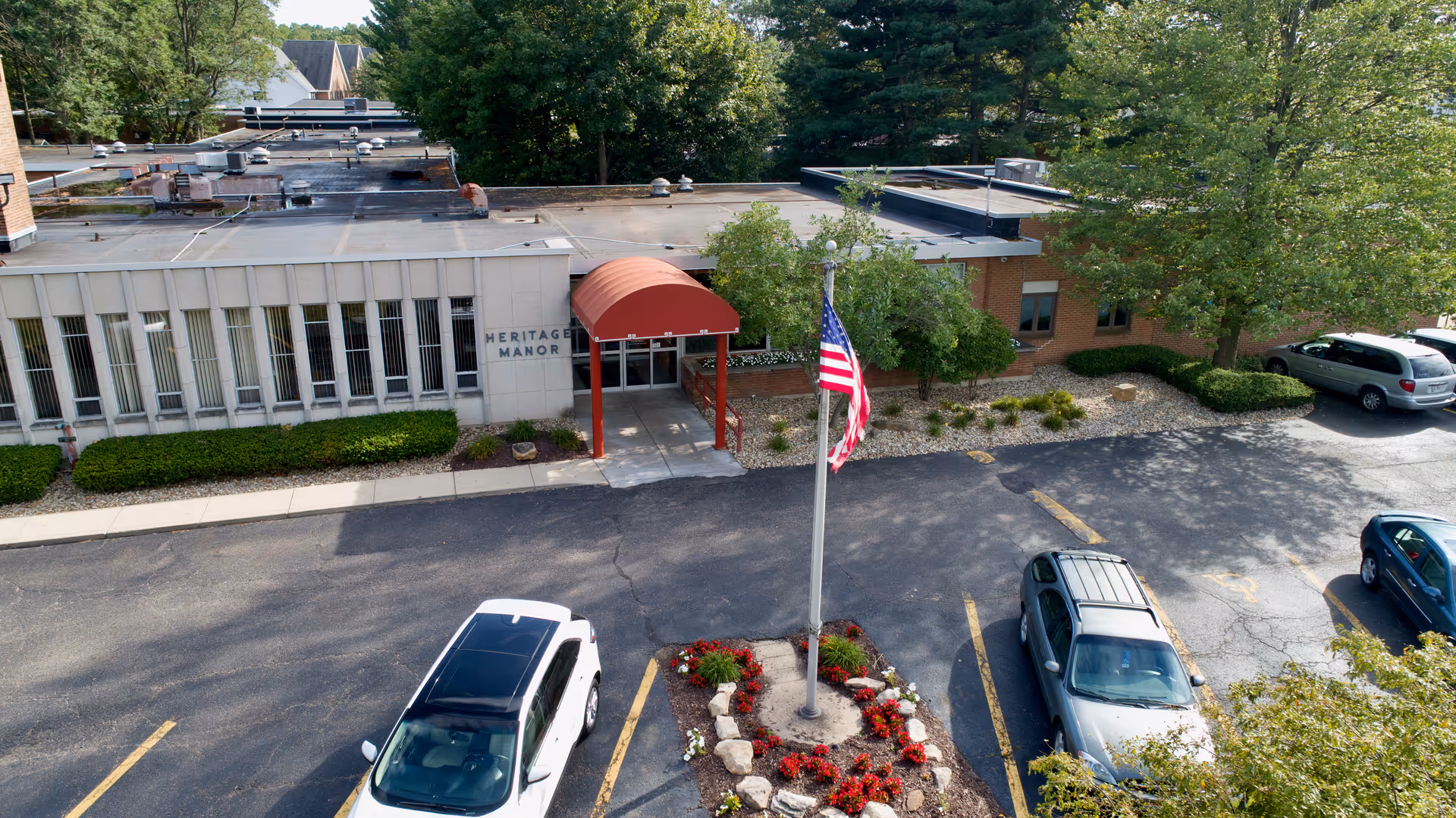 Exterior view of Heritage Manor Rehabilitation & Retirement Community building entrance with a red awning, an American flag on a flagpole surrounded by a flower bed, several parked cars, and trees in the background.