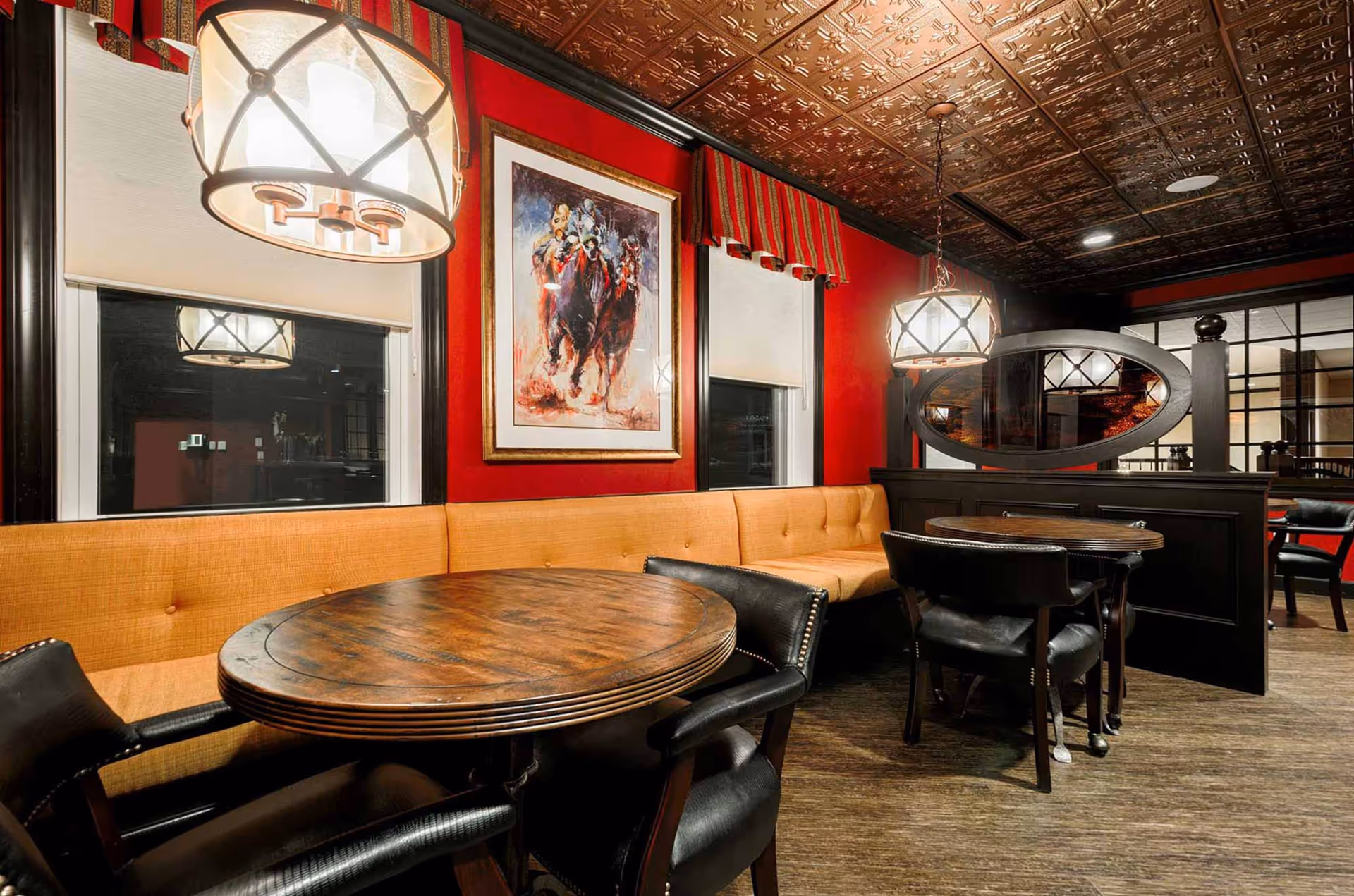 Interior dining area with round wooden tables, black leather chairs, and a long mustard-colored bench against a red wall. The ceiling has an ornate copper-colored design, and two pendant lights hang above the tables. A framed painting of horse racing is displayed on the wall between two windows with white blinds and red striped valances.