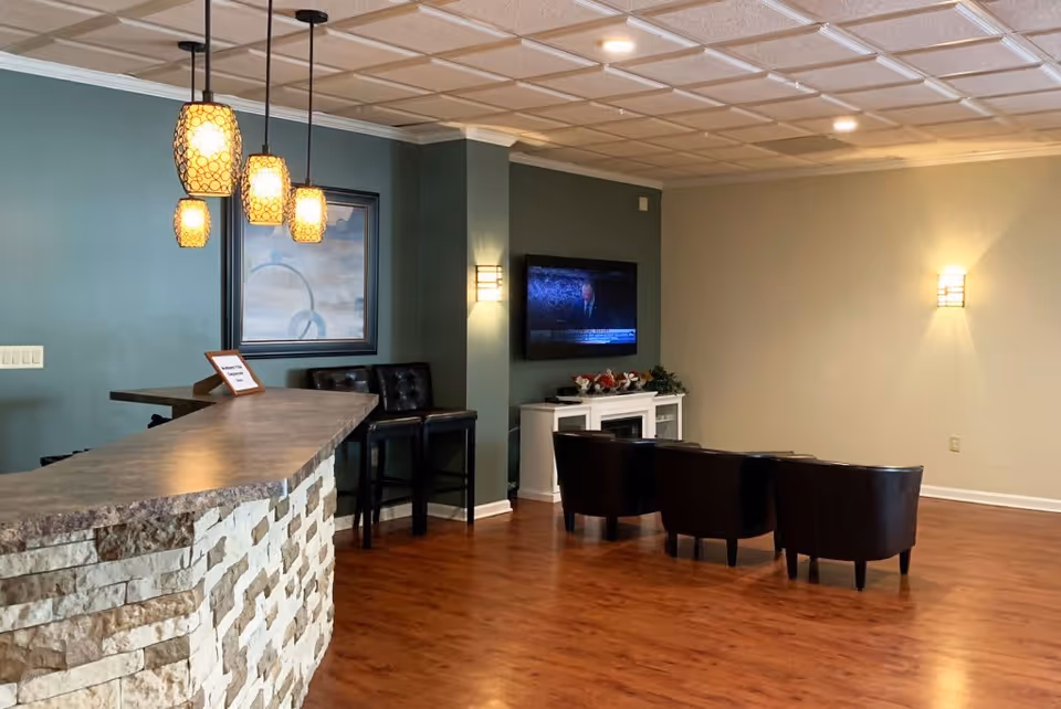 A cozy common area with a stone-faced counter and hanging pendant lights on the left, three black leather chairs facing a wall-mounted TV above a white cabinet, wooden flooring, and soft wall lighting.