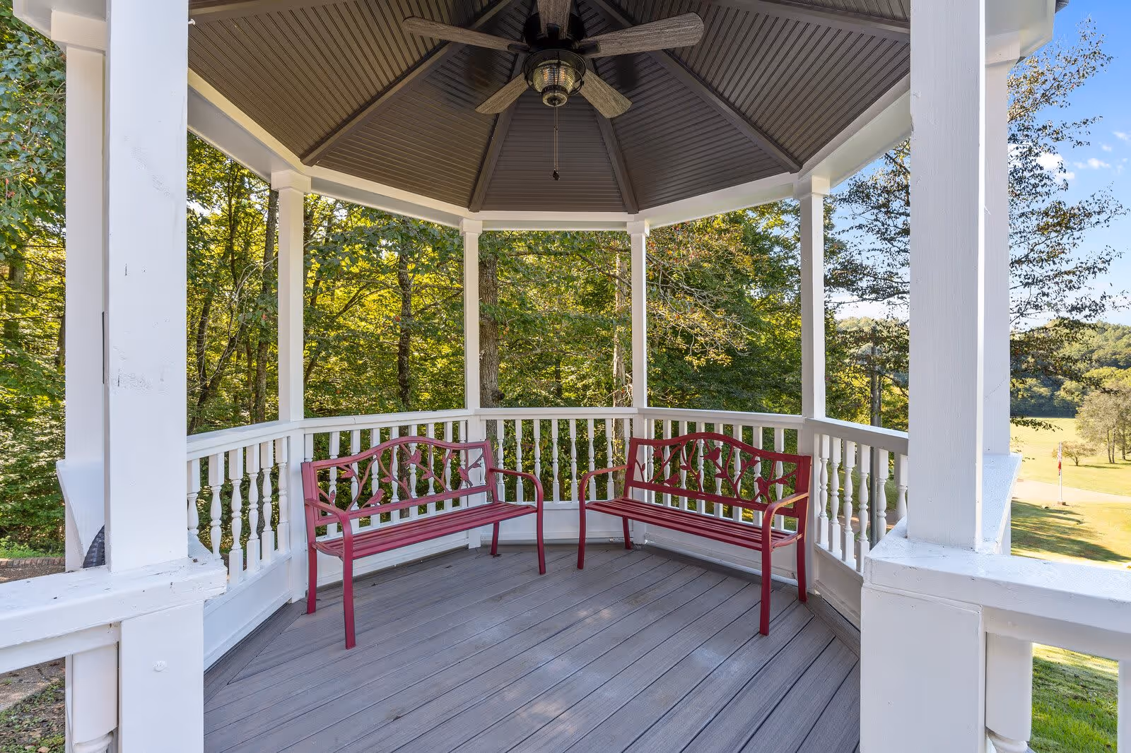 A white gazebo with a gray ceiling and ceiling fan, containing two red metal benches with decorative backs, surrounded by green trees and a grassy landscape in the background.