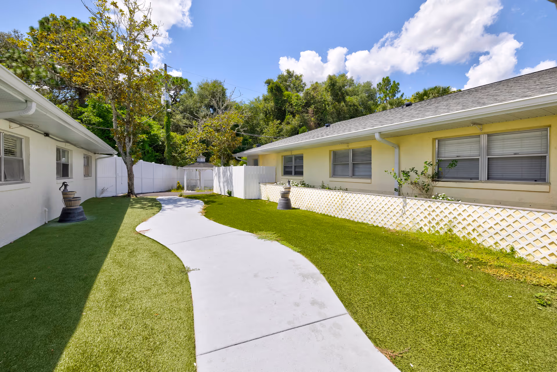Outdoor walkway at The Gardens of Eastbrooke with a curved concrete path bordered by green grass and two buildings on either side. There is a white fence and a small gazebo in the background under a blue sky with some clouds.