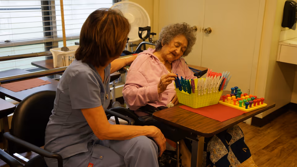 An elderly woman in a wheelchair is engaged in a therapeutic activity involving colorful pegs and clothespins on a table, while a caregiver in scrubs sits beside her offering support in a well-lit room with windows and wooden flooring.