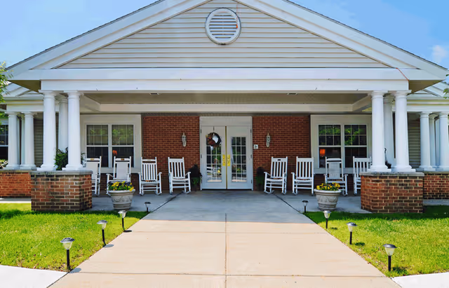 Covered front entrance of a brick building with white columns, a row of rocking chairs on the porch, potted plants, and double glass doors.