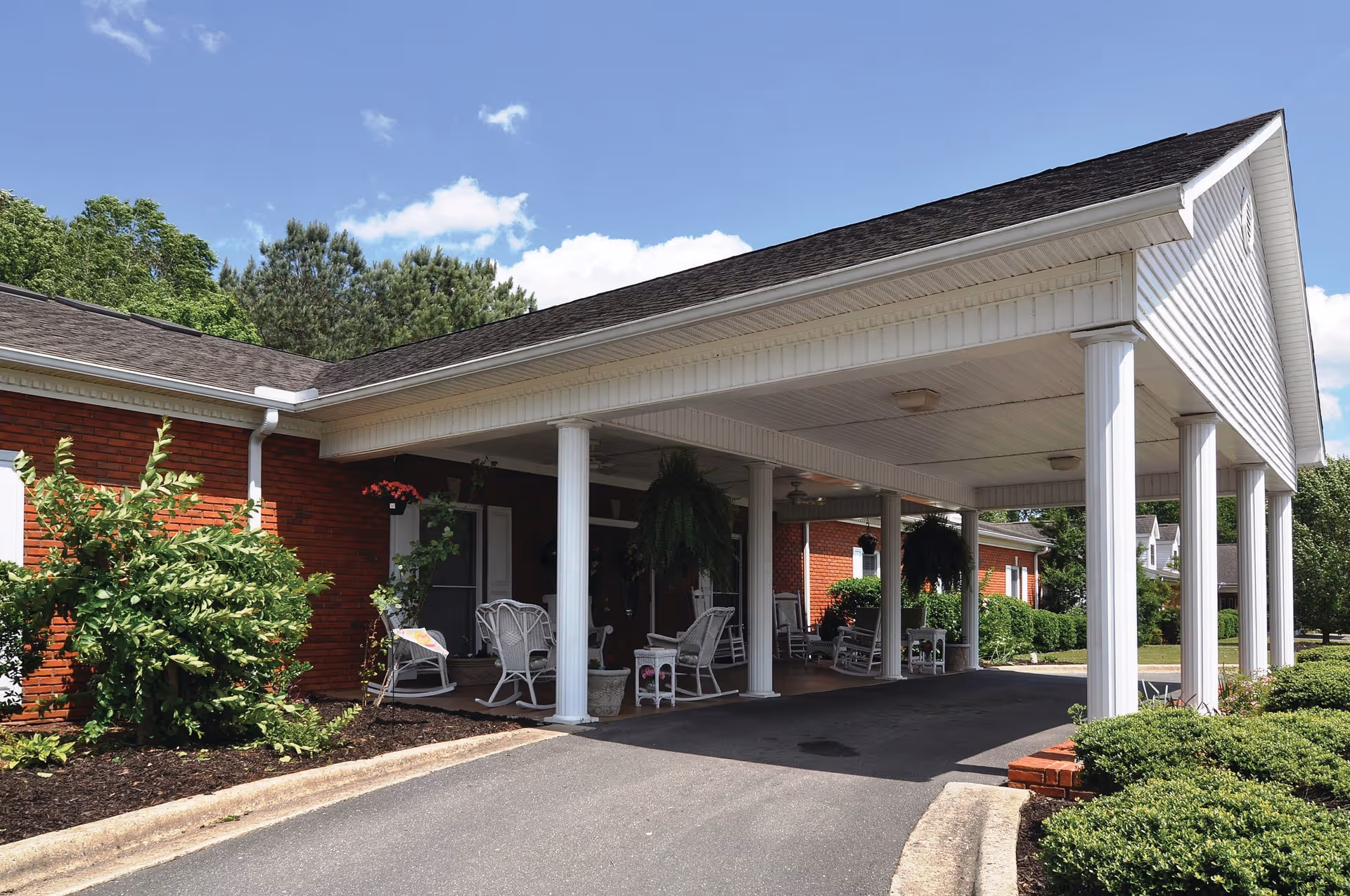 Covered entrance area of a brick building with white columns and a roof overhang. Several white rocking chairs and small tables with potted plants are arranged under the covered area. Green shrubs and trees surround the building under a blue sky with some clouds.