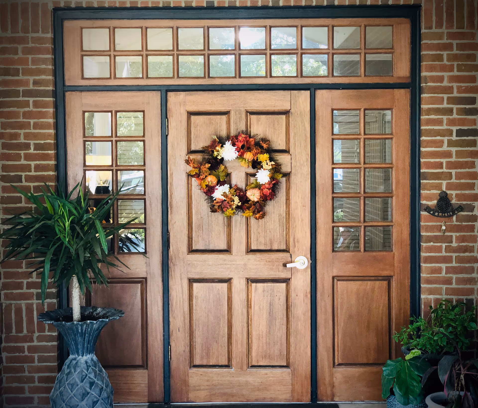 Wooden front door with glass panels on either side and above, decorated with a colorful autumn wreath. The door is set in a brick wall with potted plants on both sides.