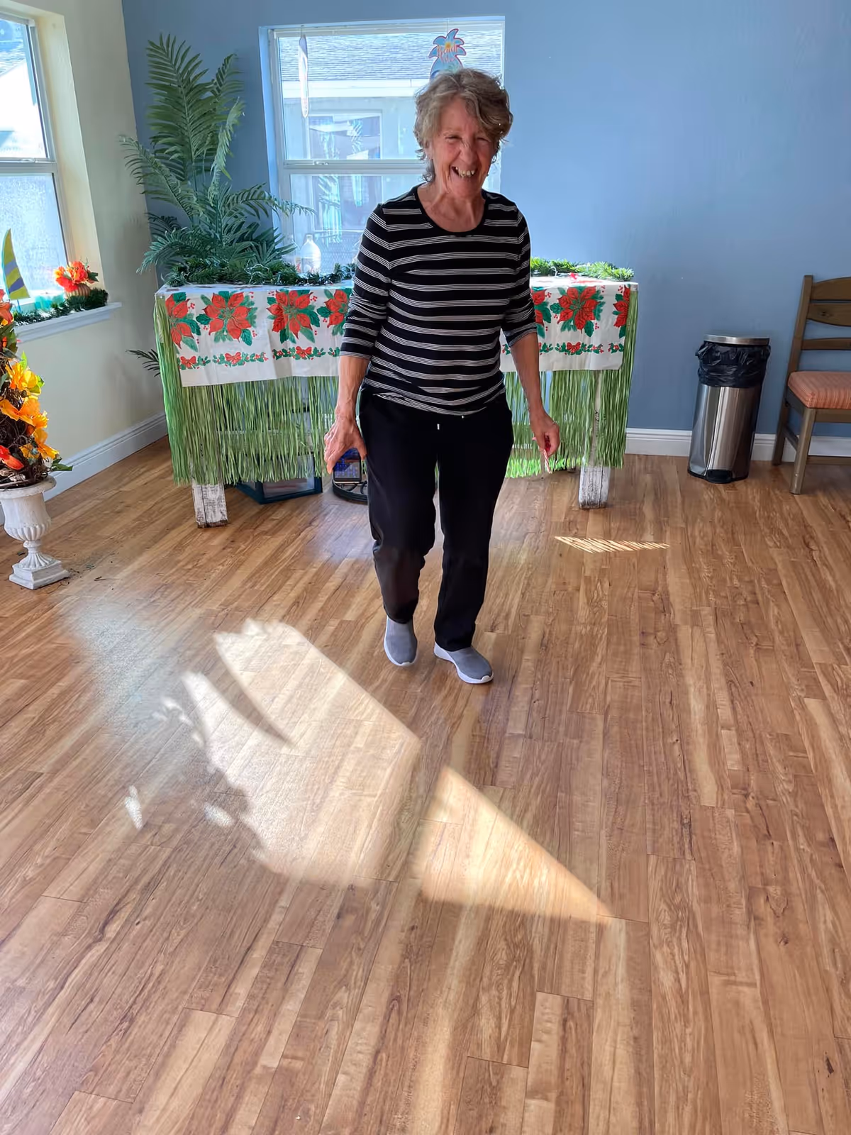 An elderly woman wearing a black and white striped shirt and black pants is smiling and walking on a wooden floor inside a room. Behind her is a table covered with a festive tablecloth decorated with red poinsettias and green fringe. The room has light blue walls, a window letting in sunlight, a potted plant, a trash can, and a chair with an orange cushion.