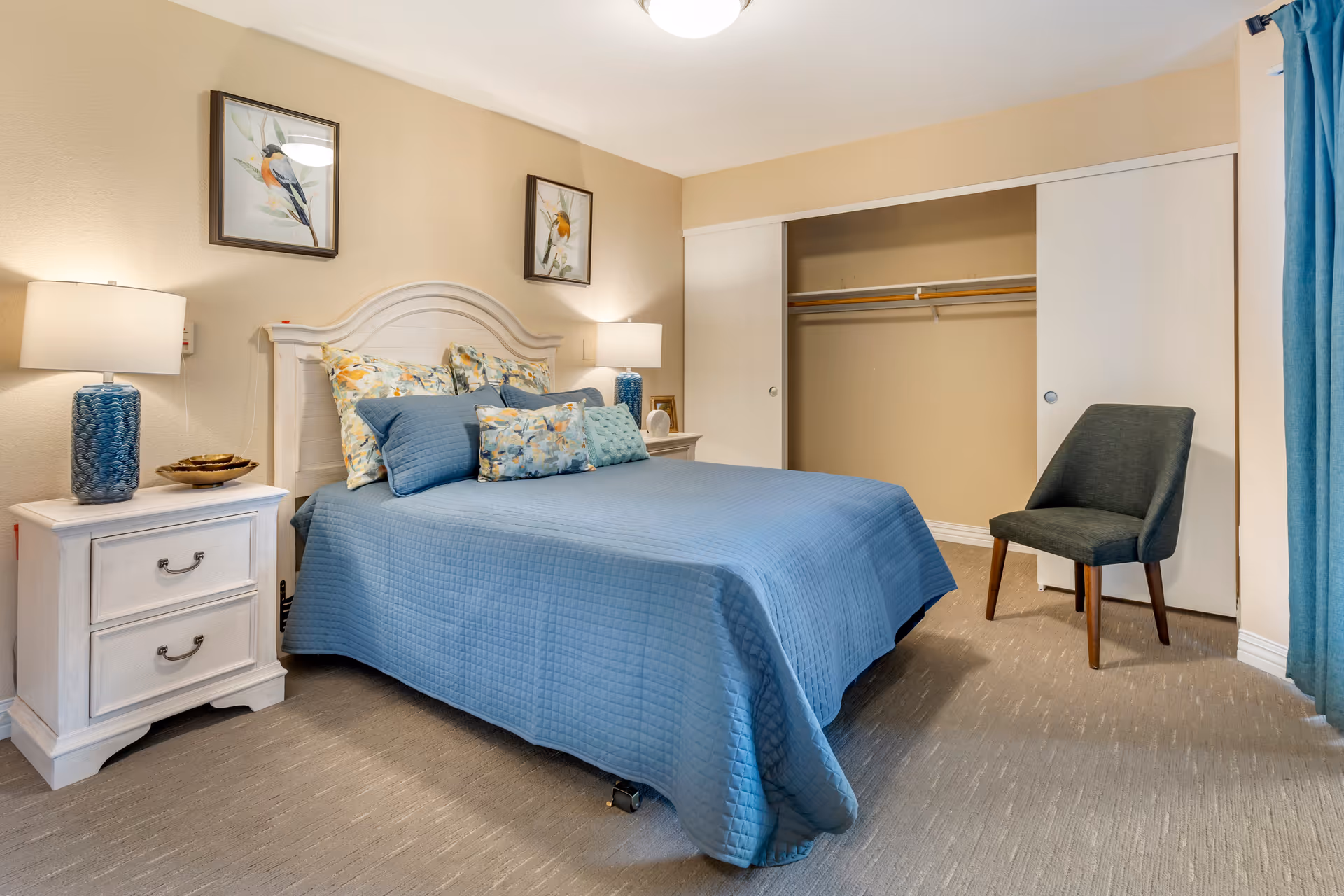 A bedroom with a bed covered in a blue quilt and multiple pillows, two white nightstands with blue lamps on each side, a dark gray chair, an open closet with sliding doors, beige walls, and two framed bird paintings above the bed.