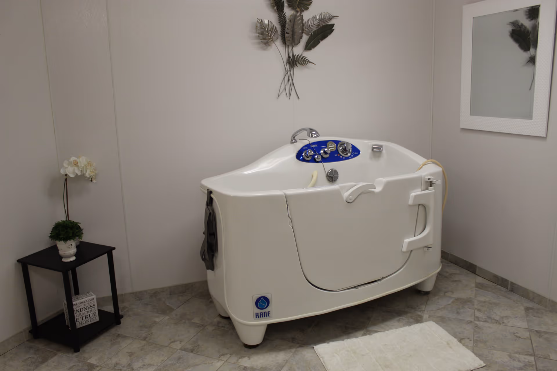A small, white walk-in bathtub with control knobs and a door, placed in a tiled room with light-colored walls. There is a black side table with a white orchid and a small green plant on it, a decorative wall hanging above the tub, a white-framed mirror on the wall, and a white bath mat on the floor.