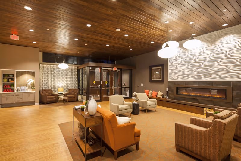 Warm, spacious senior living lounge with multiple armchairs, a fireplace, wooden ceiling, and a seating area near the entrance.