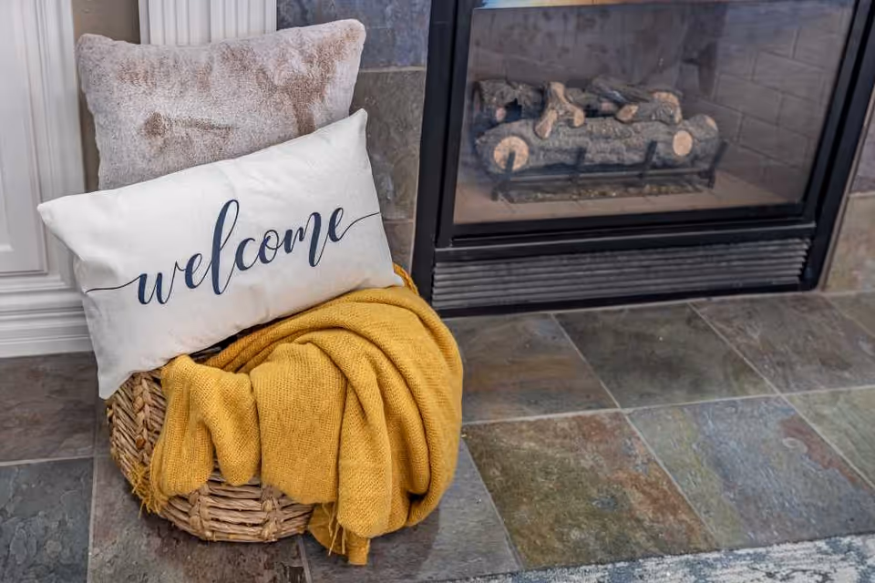 A cozy corner with a woven basket holding a yellow knitted blanket and two pillows, one of which has the word 'welcome' written on it, placed on a tiled floor next to a fireplace with logs inside.