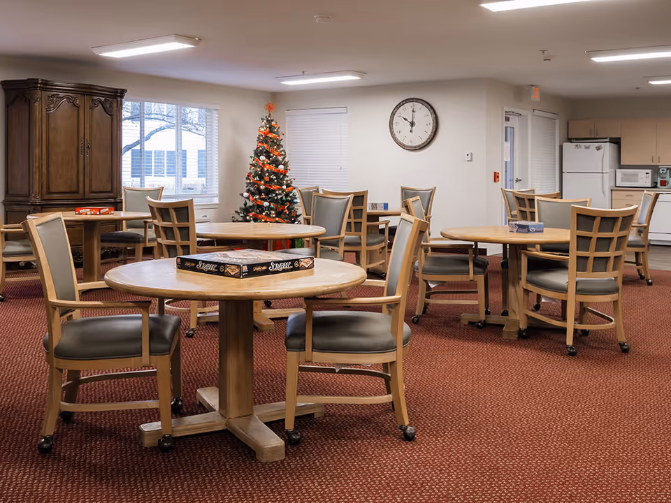 A common area in a senior living facility with several round wooden tables and chairs on a red carpet. A decorated Christmas tree is visible near the windows, and a large clock hangs on the wall. In the background, there is a kitchen area with a refrigerator, microwave, and cabinets. Board games are placed on the tables.