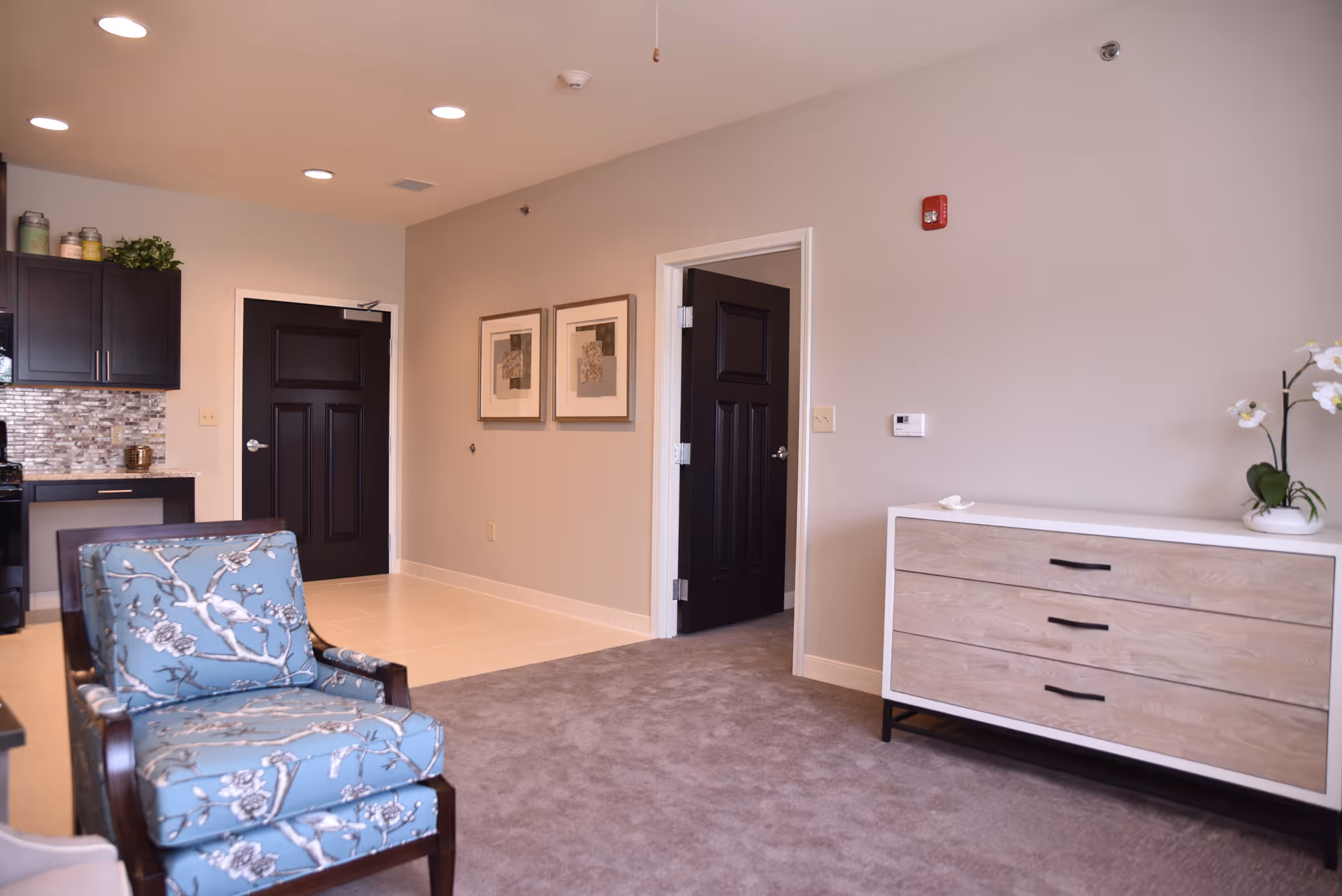 Interior view of a room in The Glen facility showing a blue patterned armchair, a wooden dresser with three drawers, two framed artworks on a beige wall, and two black doors. The room has a mix of carpet and tile flooring, with a small kitchen area visible in the background.