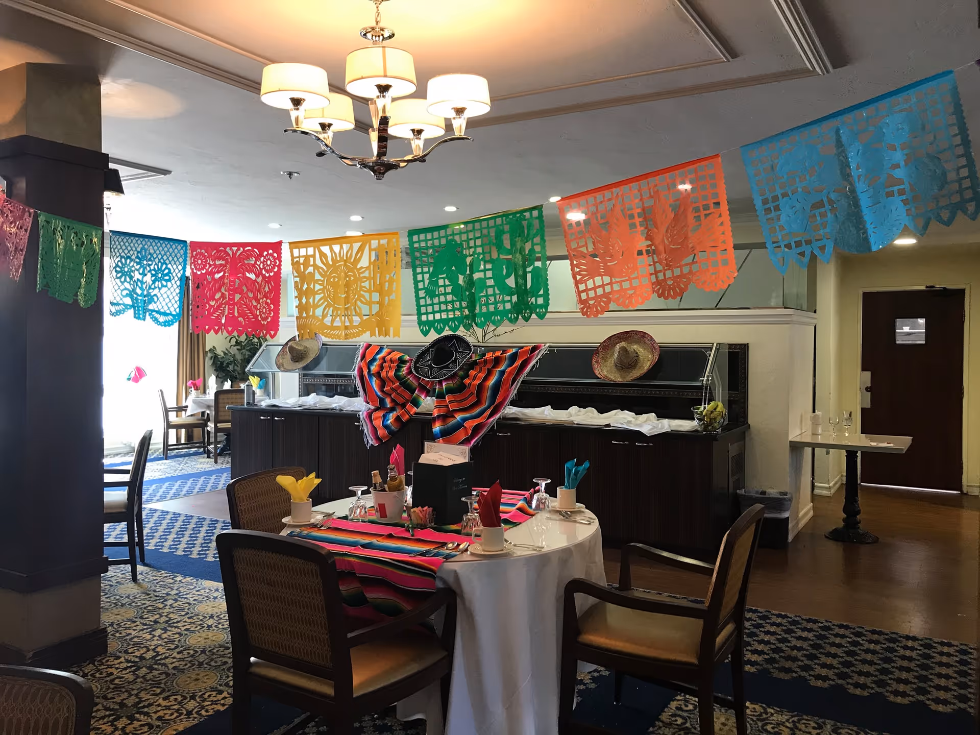 Dining area decorated with colorful papel picado banners hanging from the ceiling and sombreros on the buffet counter. A table is set with a white tablecloth, colorful striped runner, napkins, glasses, and chairs around it. The room has patterned carpet and a chandelier overhead.