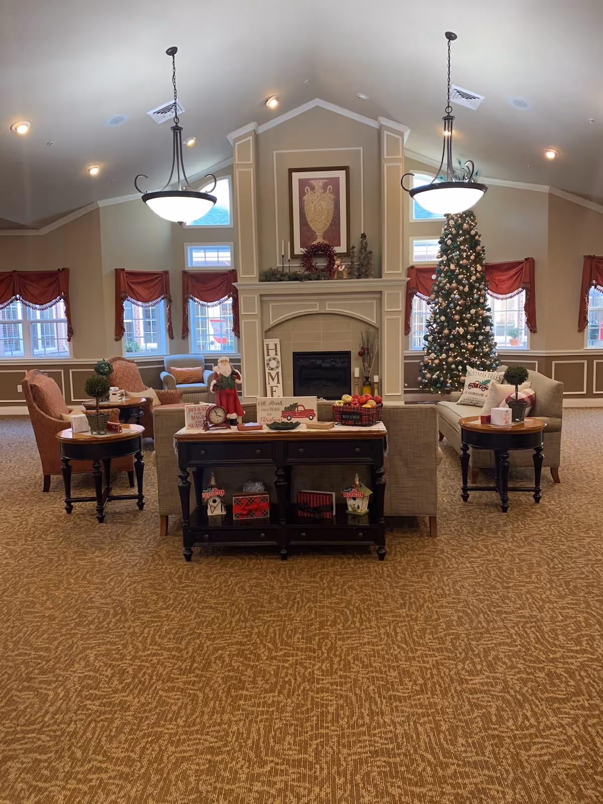 A cozy living room area decorated for Christmas with a large Christmas tree adorned with ornaments and lights. The room features a fireplace with holiday decorations on the mantel, including a wreath and candles. There are several armchairs and sofas arranged around a central table, with side tables holding small plants and holiday decor. The walls have windows with red valances, and two hanging light fixtures illuminate the space.