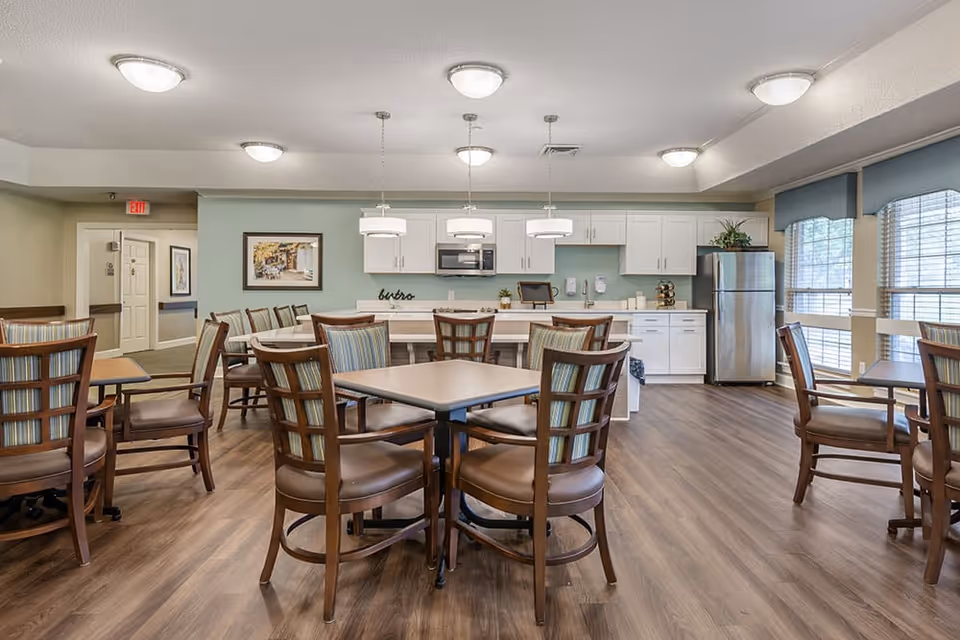 A bright and clean dining area in a senior living facility with several wooden tables and chairs arranged neatly on a wood floor. The back wall features a kitchen area with white cabinets, a stainless steel refrigerator, a microwave, and a countertop with three pendant lights hanging above. Large windows with blue valances let in natural light.