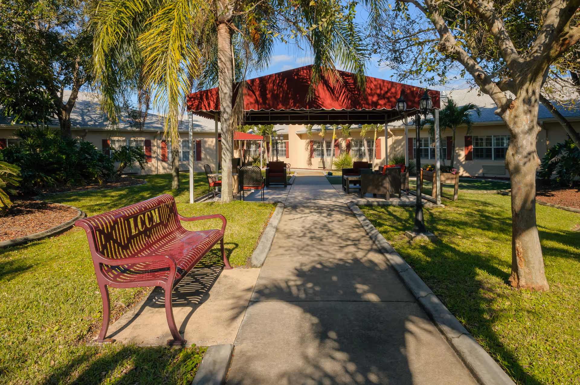 Outdoor courtyard area of a senior living facility with a concrete walkway leading to a covered seating area with chairs and tables. There are palm trees and other greenery surrounding the path, and a maroon bench with the letters 'LCCM' is positioned on the left side of the walkway. The building is visible in the background under a clear blue sky.