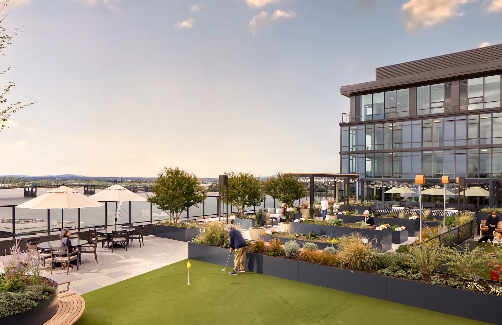 Outdoor terrace area at The Springs at The Waterfront featuring a putting green with a person practicing golf, seating areas with tables and umbrellas, landscaped garden beds, several people enjoying the space, and a modern multi-story building with large windows in the background overlooking a body of water.