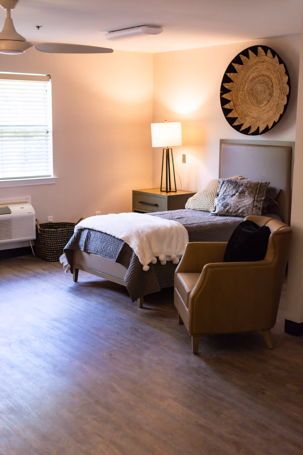 A cozy bedroom in an assisted living facility featuring a single bed with gray bedding and a white throw blanket, a bedside table with a modern lamp, a tan armchair with a black pillow, a large decorative circular wall hanging, a window with blinds, and a ceiling fan.