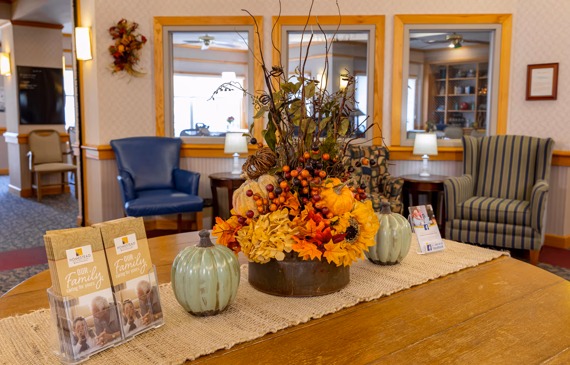 Decorated lobby table with an autumn floral centerpiece, small decorative pumpkins, brochures, and armchairs in the background.