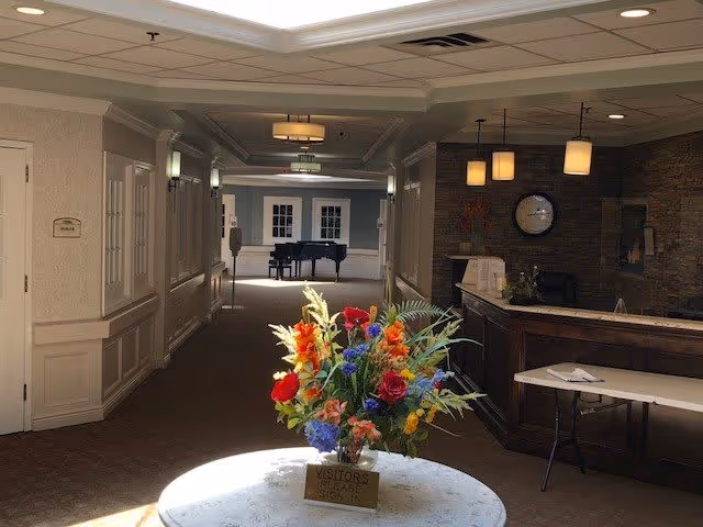 Interior view of a senior living facility hallway with a round table in the foreground holding a colorful flower arrangement and a sign that says 'Visitors Please Sign In'. To the right is a reception desk with a clock on the stone wall behind it. At the end of the hallway, a grand piano is visible in a well-lit room.