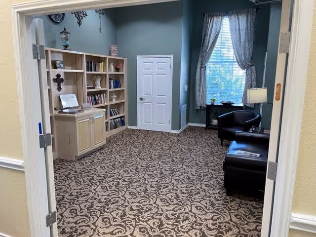Interior view of a cozy common area with patterned carpet, a bookshelf filled with books and decorative items, a small cabinet, two black armchairs, a side table with a lamp, and a window with curtains letting in natural light.