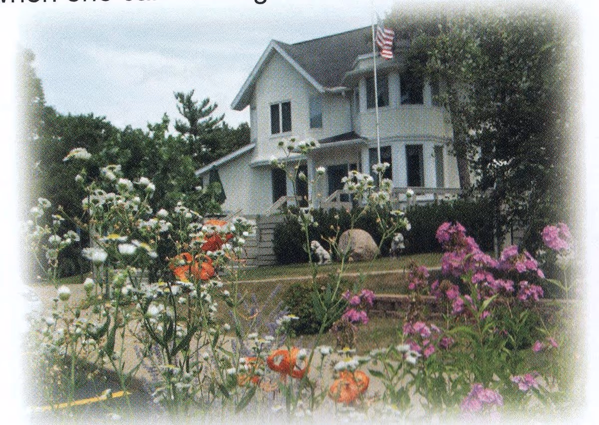 A white two-story house with a dark roof, surrounded by a garden with various flowers including white, orange, and purple blooms. An American flag is flying on a flagpole in front of the house.