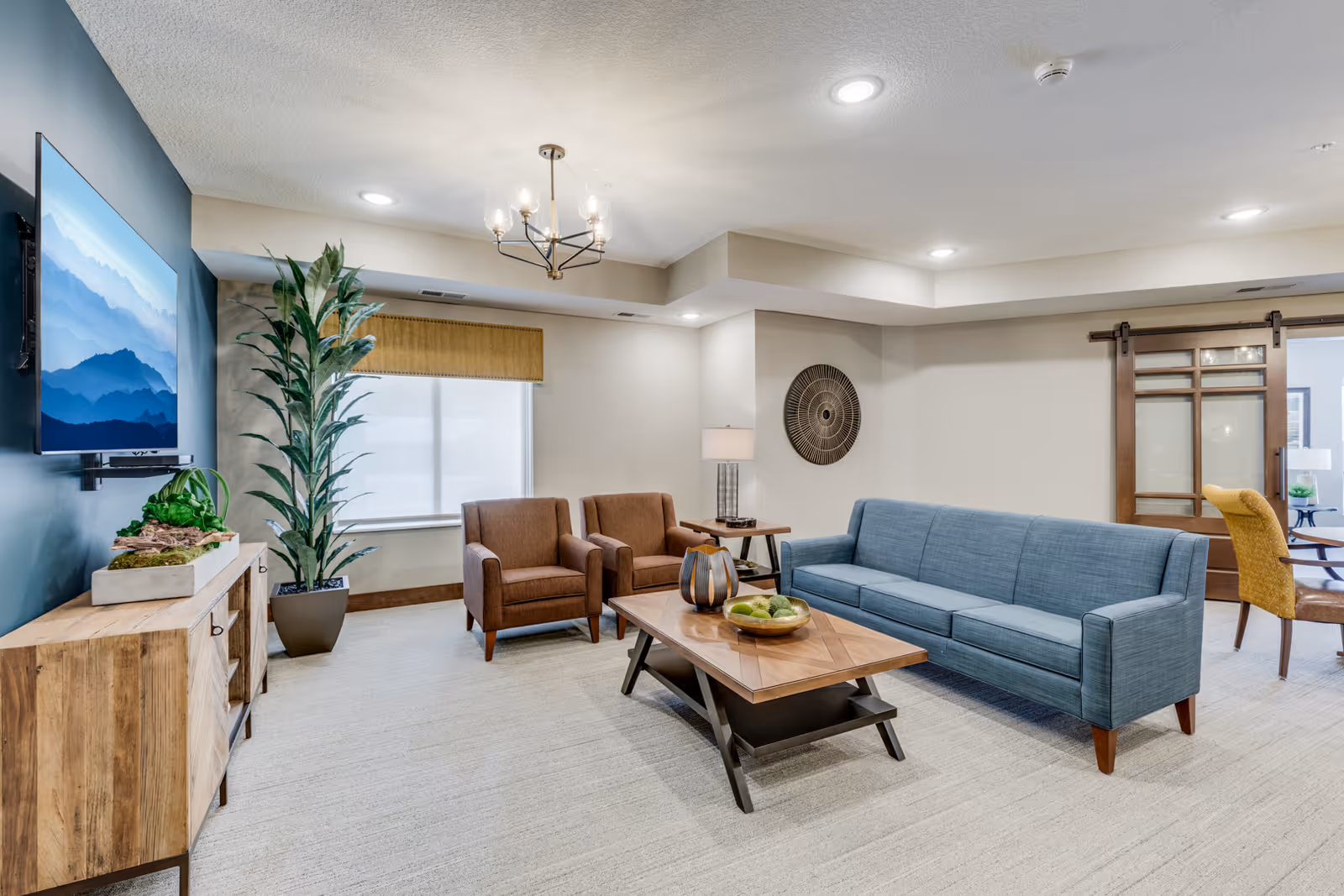 A modern living room with a blue sofa, two brown armchairs, a wooden coffee table with decorative items, a wooden media console with a flat-screen TV mounted above it, a tall potted plant, and a window with a bamboo shade. The room has beige walls, recessed lighting, a chandelier, and a sliding barn door leading to another room.