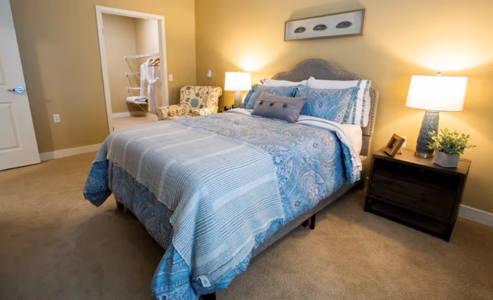 A cozy bedroom with a neatly made bed featuring blue and white patterned bedding. There is a nightstand with a lamp, a small plant, and a picture frame on the right side of the bed. A comfortable armchair with a floral pattern is placed near an open doorway leading to a walk-in closet with hanging robes and shelves. The walls are painted a warm beige color, and the room is carpeted.