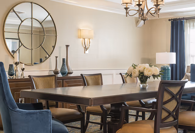 A well-lit dining room with a large rectangular wooden table surrounded by cushioned chairs. A vase with white flowers is placed on the table. The room features a decorative round mirror on the wall, a sideboard with various vases, a wall sconce, a chandelier, and a window with blue curtains allowing natural light to enter.