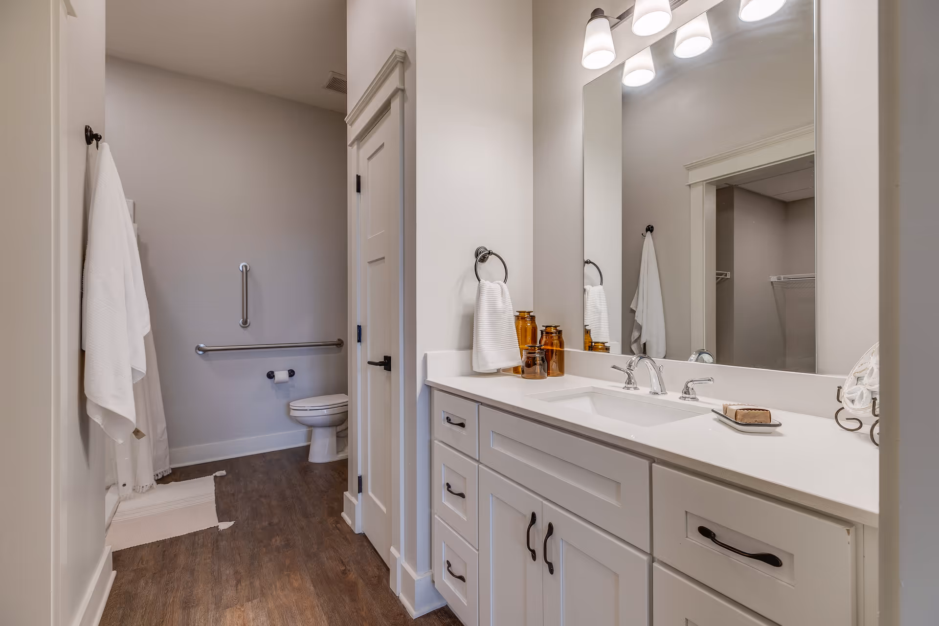 A clean and modern bathroom with white cabinetry, a large mirror, and a countertop with a sink. There are amber-colored glass containers and a soap dish on the counter. The bathroom has wood-look flooring, white walls, and a toilet with grab bars for accessibility. White towels hang on hooks and a towel ring.