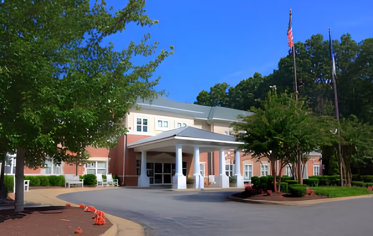Front exterior view of a senior living facility building with a covered entrance supported by white columns, surrounded by trees and greenery under a clear blue sky.