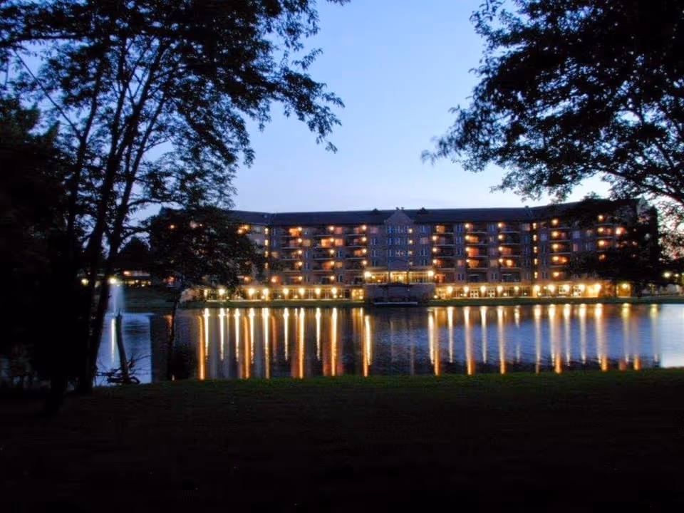 A large multi-story building illuminated at dusk, reflecting lights on a calm body of water in front. Trees frame the scene on both sides, and the sky is clear with a fading blue hue.