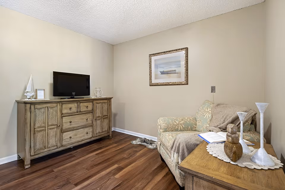 A cozy living room area with a floral patterned armchair draped with a beige throw blanket, a wooden side table with decorative items including two white candle holders and a small sculpture, a wooden cabinet with a flat-screen TV on top, and a framed picture of a boat on the wall. The room has light beige walls and wood flooring.