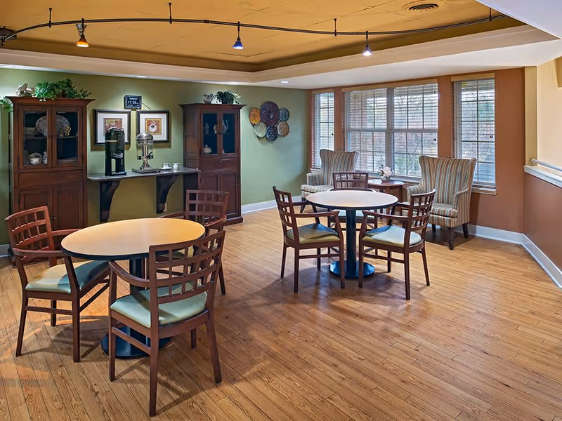 A cozy senior living common area with two round tables each surrounded by four wooden chairs with green cushions. Two striped armchairs are placed near large windows with blinds, allowing natural light to fill the room. The walls are painted in soft green and brown tones, and there are two wooden cabinets against the wall with decorative items and a coffee station between them. The ceiling has recessed lighting with small hanging spotlights.