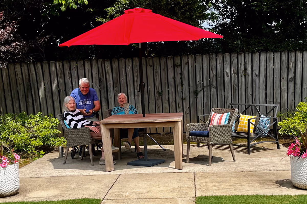Three elderly people sitting outdoors around a wooden table with a large red umbrella. They are seated on wicker chairs and a metal bench with colorful cushions. The area is paved and surrounded by greenery and a wooden fence.