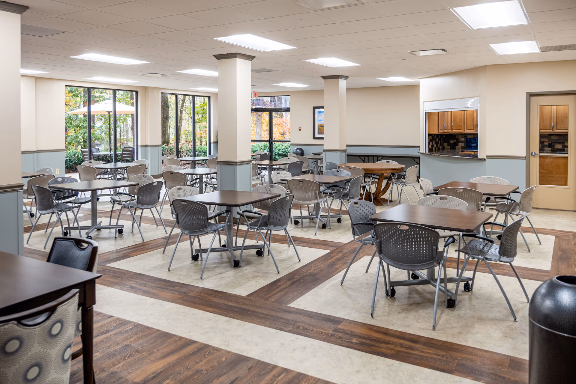A spacious dining room with multiple tables and chairs arranged neatly. Large windows on one side allow natural light to brighten the room and provide a view of outdoor greenery. The room features a combination of wood and tile flooring, beige walls with a blue-gray wainscot, and a serving window to a kitchen area. The ceiling has recessed lighting panels.