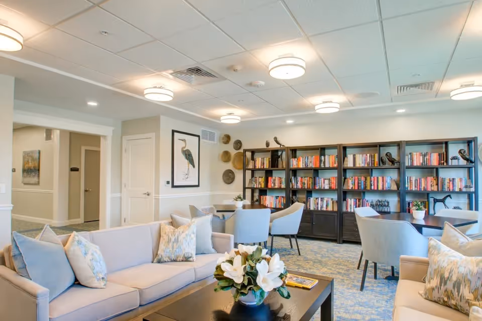 A bright and spacious living room area in a senior living facility featuring two light-colored sofas with decorative pillows, a dark wooden coffee table with a flower arrangement, several light blue armchairs around dark wooden tables, and a large bookshelf filled with books and decorative items against the back wall. The room has a patterned carpet, white walls with framed artwork, and ceiling lights providing ample illumination.