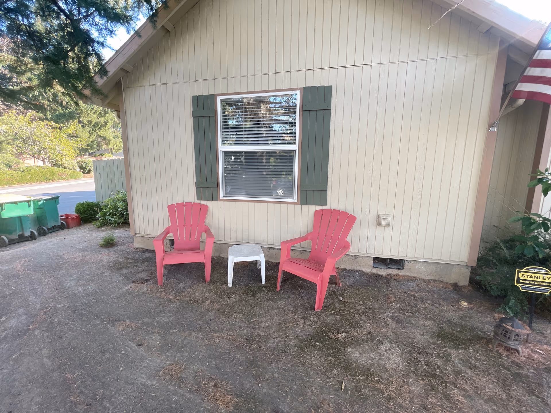 Two red outdoor chairs and a small white table sit outside a single-story house beneath a window with green shutters.