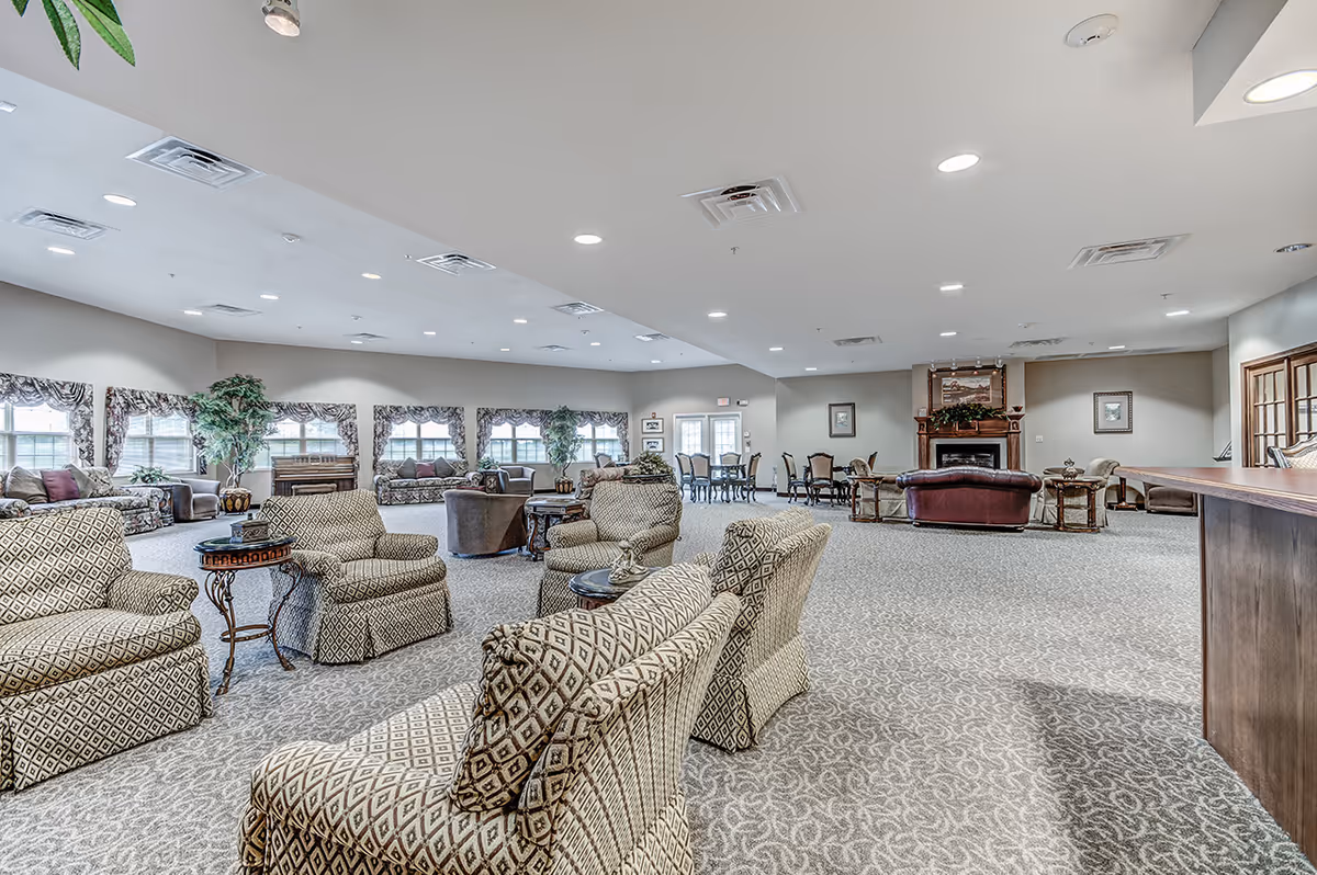 Spacious carpeted common lounge area with patterned armchairs and sofas arranged around tables and a fireplace in the background.