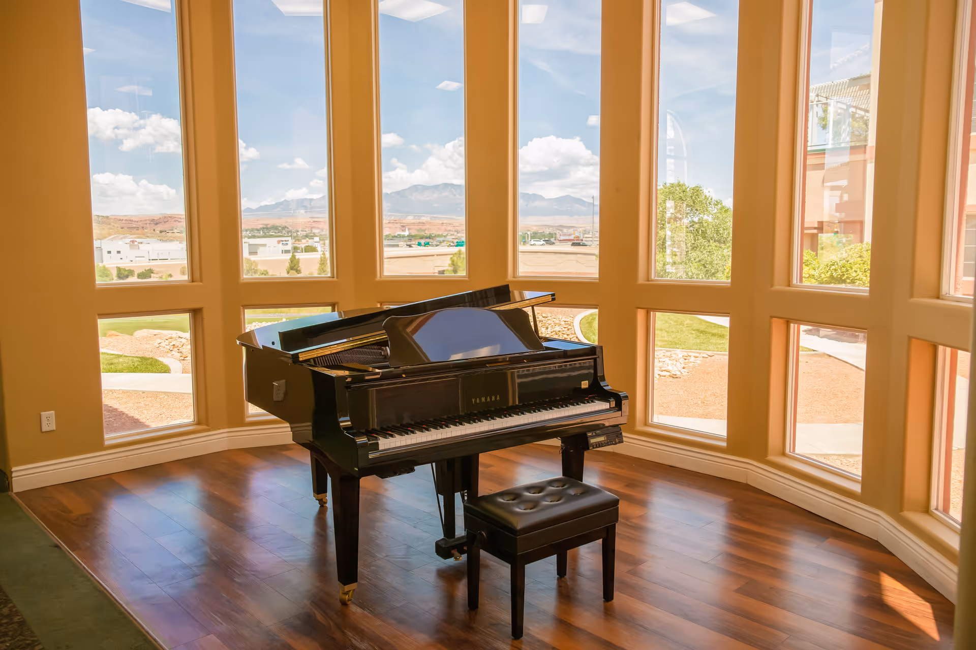 A black Yamaha grand piano with a matching bench sits on a polished wooden floor in a bright room with large, tall windows offering a view of an outdoor landscape with mountains and blue sky.