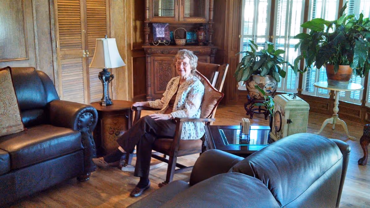 An elderly woman sitting in a wooden armchair in a cozy living room with leather sofas, a side table with a lamp, wooden furniture, and several potted plants near large windows with shutters.
