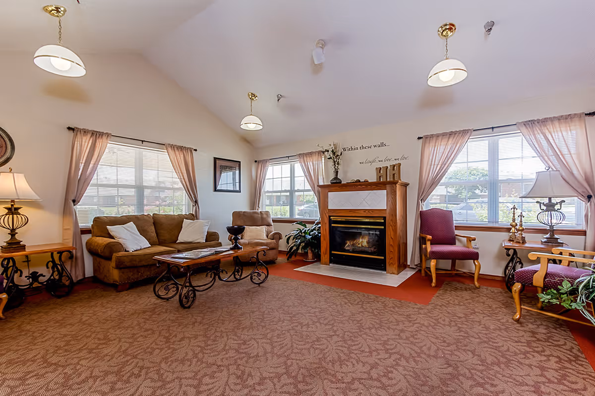A cozy senior living common area with a brown sofa, armchair, two purple chairs, and a wooden coffee table with ornate metal legs. The room features a fireplace with a wooden mantle, two large windows with sheer pink curtains, and several lamps providing warm lighting. The walls are light-colored, and the carpet has a patterned design. Above the fireplace, there is a decorative sign that reads, 'Within these walls... we laugh, we love, we live.'