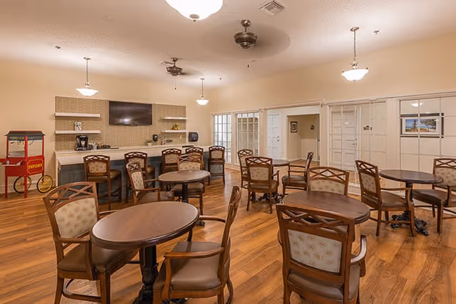 A spacious common area with several round wooden tables and cushioned chairs arranged on a wooden floor. The room features a counter with bar stools, a mounted flat-screen TV, and a popcorn machine in the corner. The walls are light-colored with multiple doors and windows, and ceiling fans and hanging light fixtures are visible.