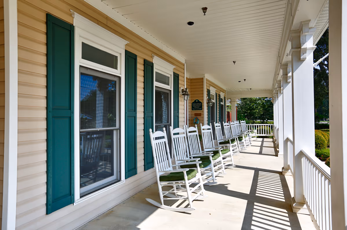 Covered front porch with a row of white rocking chairs along a tan building with green shutters.