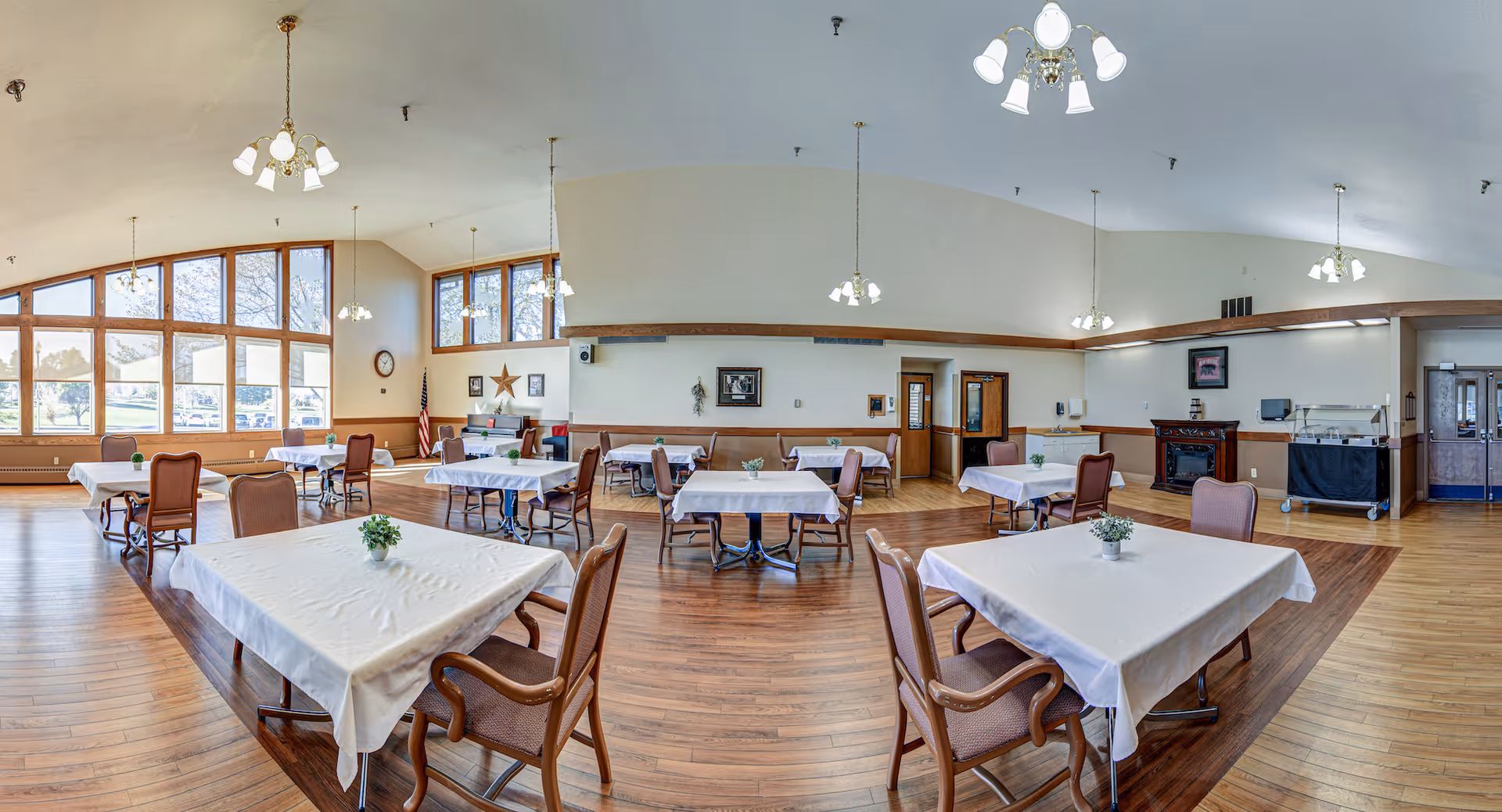 Spacious dining room with multiple tables covered in white tablecloths, each with a small green plant centerpiece. The room features large windows allowing natural light to fill the space, wooden flooring, and several hanging light fixtures. There are chairs arranged around the tables and some wall decorations including a clock, an American flag, and framed pictures.