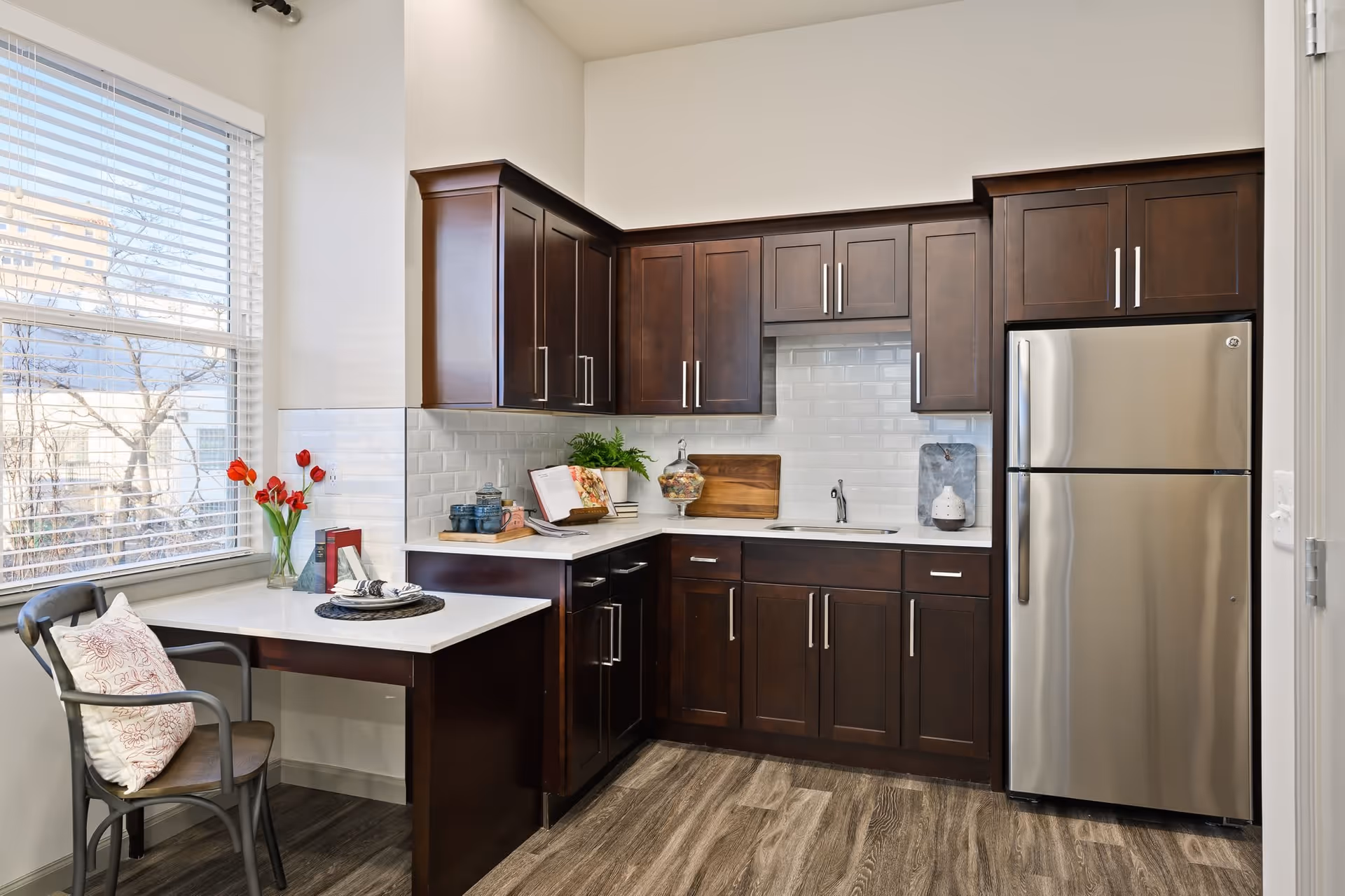 A modern kitchen with dark wooden cabinets, white countertops, and a stainless steel refrigerator. There is a small dining area with a chair and a table set near a window with blinds. The kitchen has a white tiled backsplash and wood-patterned flooring.