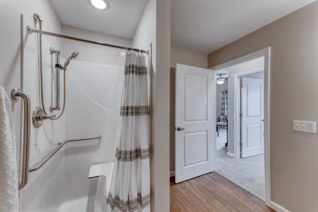 A bathroom with a walk-in shower featuring grab bars and a shower seat, a shower curtain with decorative stripes, and an adjacent hallway leading to a room with open doors and carpeted flooring.