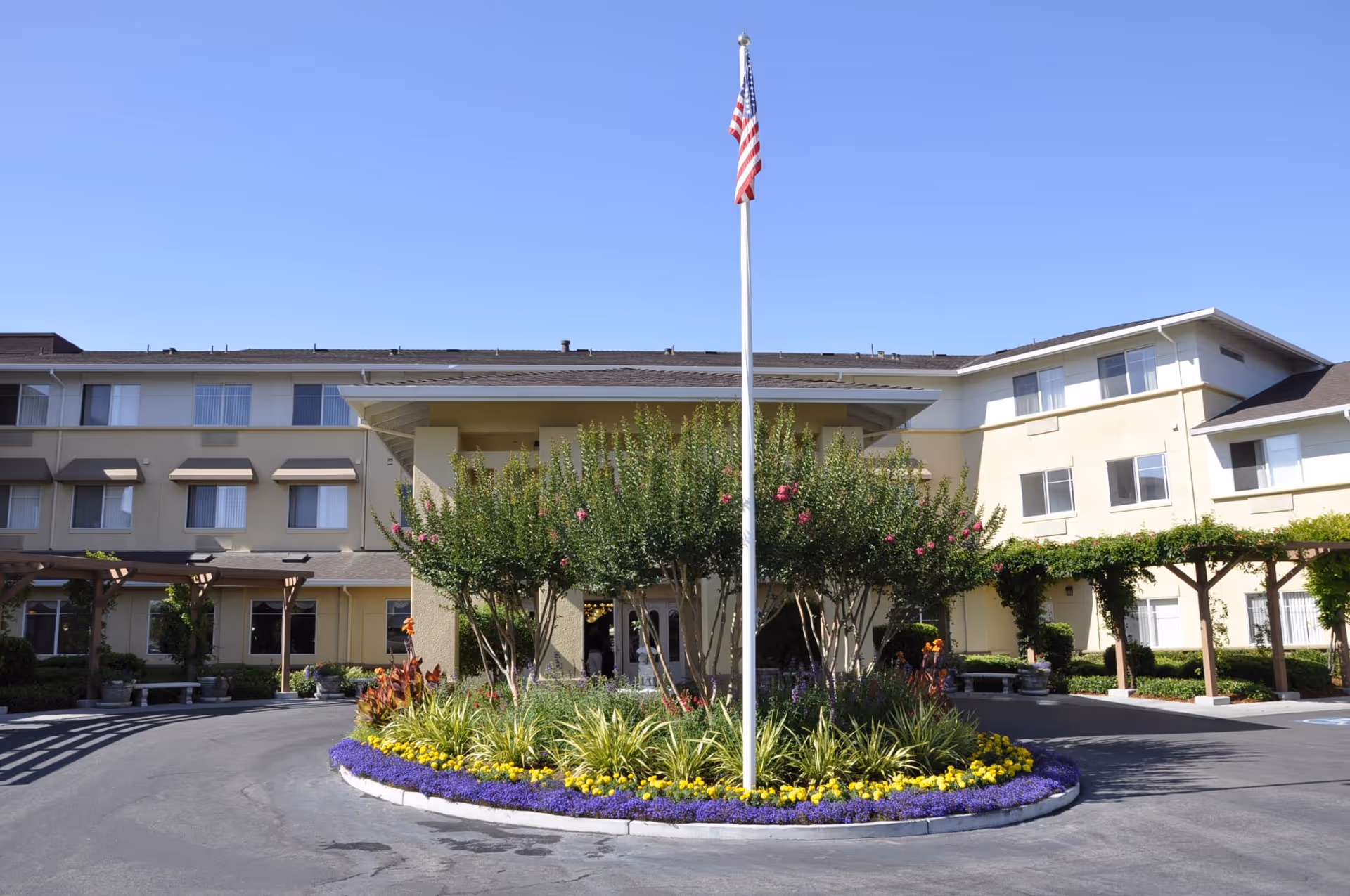 Front exterior view of Carlton Senior Living San Jose building with a circular driveway, landscaped garden with colorful flowers and shrubs, and an American flag on a flagpole in the center.