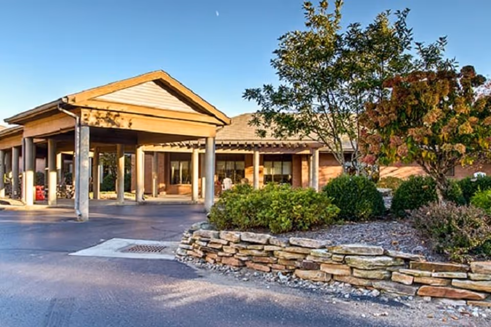 Exterior view of Elison Assisted Living & Memory Care of Marietta showing the entrance with a covered drop-off area, landscaped garden with bushes and trees, and a clear blue sky.